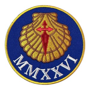 Puede incluir: Parche bordado circular con una concha dorada y una cruz roja sobre fondo azul. El parche tiene un borde dorado y el número romano "MMXXVI" en blanco. Probablemente un símbolo de peregrinación.