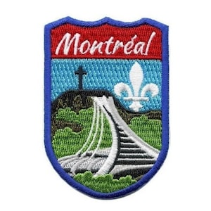 Op de afbeelding: Een geborduurde patch met een blauwe rand, een rode achtergrond met het woord "Montreal" in wit en een witte fleur-de-lis op een blauwe lucht achtergrond. De patch toont een wit gebouw met een groene heuvel en een kruis op de achtergrond.