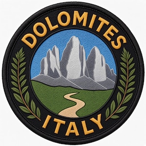 May include: A circular embroidered patch featuring a mountain range in white and grey against a blue sky. A winding tan path leads through a green landscape. The black border contains the yellow text DOLOMITES at the top and ITALY at the bottom.