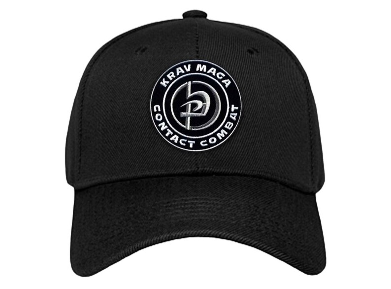 K&ouml;nnte beinhalten: Schwarze Baseballkappe mit einem wei&szlig;en gestickten Logo, auf dem "KRAV MAGA CONTACT COMBAT" mit einem stilisierten Symbol in der Mitte steht.