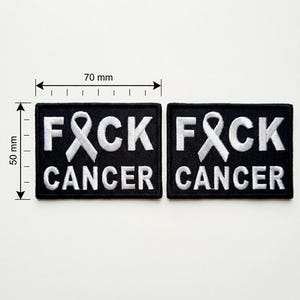 Może przedstawiać: Dwie czarne, haftowane naszywki, każda o wymiarach 7 cm na 5 cm. Naszywki zawierają biały napis "F*CK CANCER", z białą wstążką zastępującą literę "A". Naszywki są przeznaczone do przyszywania lub naprasowywania.
