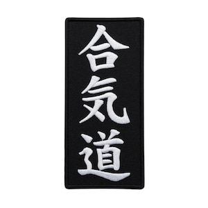 Op de afbeelding: Zwart rechthoekig embleem met witte geborduurde Japanse karakters. De karakters luiden verticaal "Aikido". Het embleem heeft een zwarte rand en staat tegen een witte achtergrond.