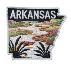 Op de afbeelding: Geborduurde patch in de vorm van Arkansas, met een landschap met een rivier en vegetatie. Het woord "ARKANSAS" is in witte blokletters geborduurd op een zwarte achtergrond. De patch heeft een witte rand.