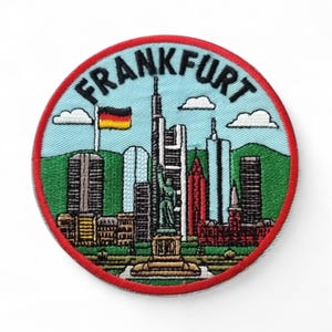 Könnte beinhalten: Runder, gestickter Aufnäher mit dem Wort "FRANKFURT" in Schwarz. Das Design zeigt eine Stadtlandschaft mit Wolkenkratzern, einer Statue und der deutschen Flagge. Er hat einen hellblauen Hintergrund, grüne Hügel, weiße Wolken und einen roten Rand. Ein Souvenirartikel.