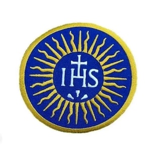 Könnte beinhalten: Ein blauer und goldener gestickter Aufnäher mit den Buchstaben "IHS" und einem Kreuz in einem Kreis aus Sonnenstrahlen.