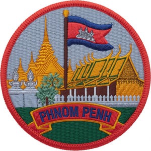 May include: Embroidered patch featuring a circular design with a red border. The patch depicts a golden temple, a red and blue flag, and the text "PHNOM PENH" on a red banner. The background is light blue.
