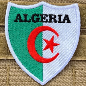 Peut inclure: Un écusson brodé blanc et vert avec le mot "ALGERIE" en lettres noires et un croissant de lune rouge et une étoile au centre.
