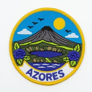 Könnte beinhalten: Gestickter Aufnäher mit einer Landschaft mit Vulkan, See und blauen Blumen. Der Aufnäher hat einen blauen Himmel mit gelber Sonne und Vögeln. Das Wort "AZORES" ist auf einem weißen Banner abgebildet.