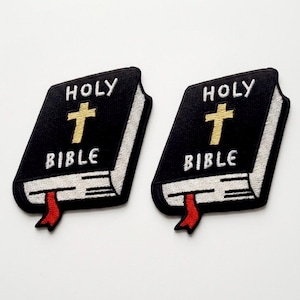 Könnte beinhalten: Zwei schwarze, gestickte Aufnäher in Buchform, jeweils mit den weißen Worten "HOLY BIBLE" und einem goldenen Kreuz. Die Aufnäher haben ein rotes Lesezeichen und weiße Nähte an den Rändern.