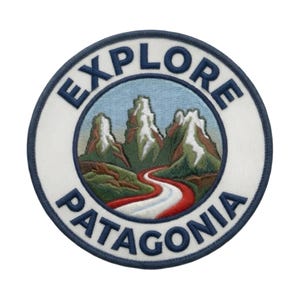 Op de afbeelding: Een ronde geborduurde patch met een witte achtergrond en een donkerblauwe rand. Het midden toont een bergketen met besneeuwde toppen, groene heuvels en een kronkelend rood-wit pad. De tekst EXPLORE PATAGONIA is in het blauw geborduurd.