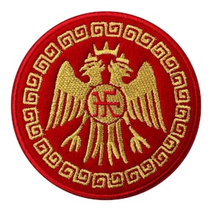 Könnte beinhalten: Ein runder, roter, gestickter Aufnäher mit einem goldenen Doppeladler-Emblem. Das Emblem ist von einem goldenen griechischen Schlüsselmuster umgeben. Über den Köpfen des Adlers befindet sich eine Krone und ein zentrales Symbol.