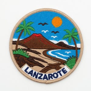 Könnte beinhalten: Gestickter Aufnäher mit einer Landschaft von Lanzarote. Das Design umfasst einen Vulkan mit roten Akzenten, Palmen, einen blauen Ozean und eine leuchtend orangefarbene Sonne. Das Wort "LANZAROTE" ist in Weiß auf blauem Grund gestickt.