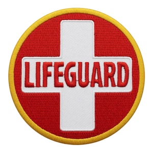 May include: Embroidered patch featuring a red cross on a white background, with the word "LIFEGUARD" in red letters. The patch is circular, with a yellow border. Ideal for lifeguard apparel.