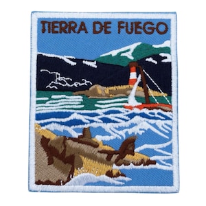 May include: Embroidered patch featuring a coastal scene with the text "TIERRA DE FUEGO." The design includes a lighthouse, sailboat, mountains, and ocean waves in shades of blue, brown, and white, with a white border.