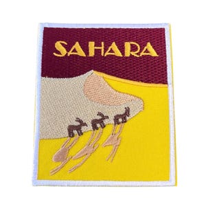 Op de afbeelding: Een geborduurde patch met een gele achtergrond en een witte rand. De patch toont een bruine kamelenkaravaan die een zandduin oploopt. Het woord "SAHARA" is geborduurd in geel op een kastanjebruine achtergrond bovenaan de patch.