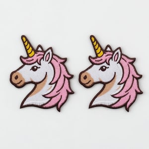 Könnte beinhalten: Zwei gestickte Einhorn-Aufnäher. Jeder Aufnäher zeigt einen weißen Einhornkopf mit rosa Mähne, gelbem Horn und brauner Umrandung. Die Aufnäher sind für das Aufnähen auf Stoff konzipiert.