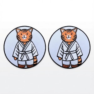 May include: Two circular embroidered patches featuring an orange tabby cat with dark brown stripes. The cat is wearing a white martial arts uniform with a matching white belt. Each patch has a light blue background and a thick black border.