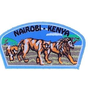 Può includere: Una toppa ricamata blu e bianca con il testo "NAIROBI - KENYA" e un'immagine di tre leoni in un ambiente di savana.