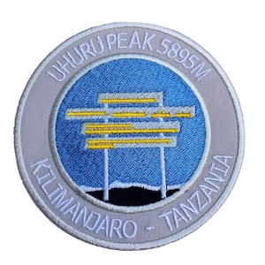 Könnte beinhalten: Ein weißer gestickter Aufnäher mit blauem Hintergrund und einer gelben Silhouette des Kilimandscharo. Der Text "Uhuru Peak 5895m Kilimanjaro - Tansania" ist um den Rand des Aufnähers gestickt.