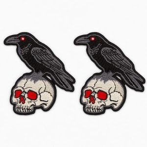 May include: Two embroidered patches featuring a black raven perched atop a skull. The raven has red eyes and detailed feathering. The skull is off-white with red eye sockets. These patches are suitable for jackets, bags, or other fabric items.