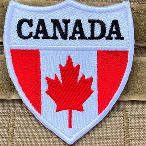 Op de afbeelding: Witte geborduurde patch met een rode Canadese vlag en het woord "CANADA" in zwarte letters.