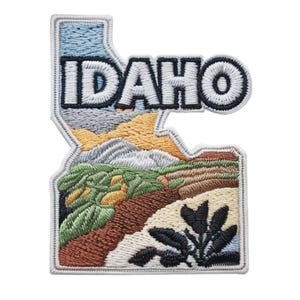 Könnte beinhalten: Gestickter Aufnäher in Form von Idaho, mit dem Staatsnamen in weißen Buchstaben. Der Aufnäher zeigt eine Landschaft mit Bergen, Feldern und einem Fluss, mit Farben wie Blau, Grün, Braun und Gelb.