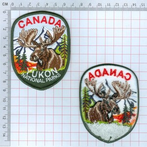 May include: Two embroidered patches featuring a brown moose, mountains, and trees. The larger patch reads CANADA YUKON NATIONAL PARKS. The smaller patch reads CANADA. A ruler shows the larger patch is approximately 9 cm tall.