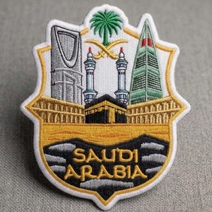 Può includere: Toppa ricamata con un design a scudo raffigurante monumenti iconici dell'Arabia Saudita. La toppa include la scritta "SAUDI ARABIA" in oro, insieme a immagini di una palma, edifici e una moschea. La toppa ha un bordo dorato, nero e bianco.