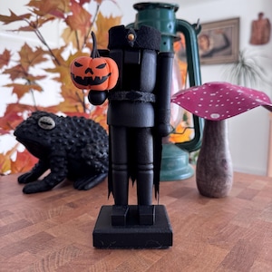 May include: A black wooden nutcracker figure standing on a square base, wearing a black hat with a gold stud. The figure holds a small orange pumpkin with a black painted jack-o'-lantern face in its right hand and wears a black cape-like garment.