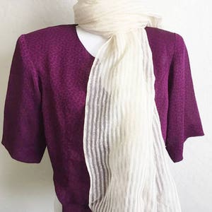 May include: A plum-colored blouse with a subtle spotted pattern and short sleeves is paired with a cream-colored, vertically striped scarf. The scarf is draped over the blouse, creating a layered look. The clothing is displayed on a white mannequin.
