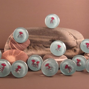 May include: A collection of clear glass marbles with a light blue hue, each featuring a red jellyfish design. The marbles are arranged on and around various stones, creating a unique display. The marbles are approximately 1 inch in diameter.