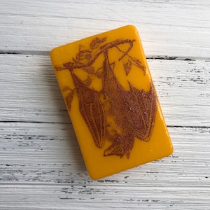 May include: A yellow rectangular soap bar with a brown silhouette of a bat hanging upside down from a branch. The bat has detailed wings and a pointed nose.