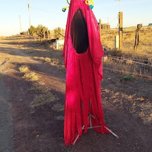 May include: A red, fuzzy costume with a large, black mouth and two large, white eyes. The costume is on a tripod stand and is set up outdoors.
