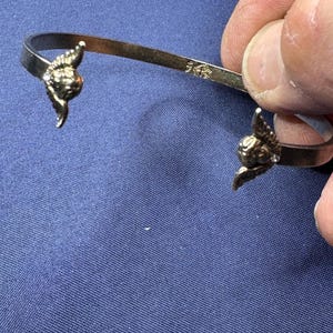 May include: A gold-colored cuff bracelet with decorative winged accents at each end. The bracelet is held against a blue background. The metal is stamped with "14K".