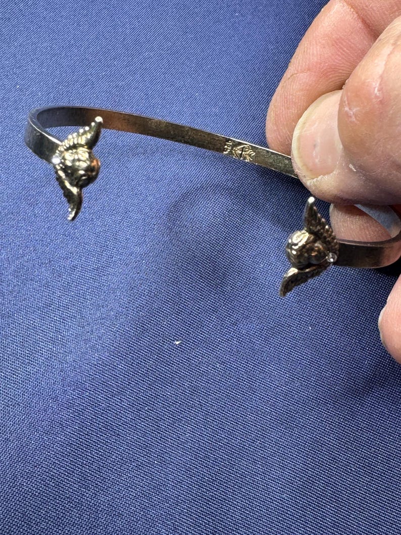May include: A gold-colored cuff bracelet with decorative winged accents at each end. The bracelet is held against a blue background. The metal is stamped with "14K".