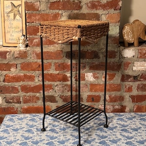 May include: A decorative plant stand with a woven top and a black metal frame. The stand has a lower shelf and is placed on a blue and white patterned tablecloth. The background features a brick wall and a framed artwork.