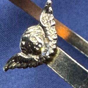 May include: Close-up of a silver-toned compass with an ornate, detailed design. The compass features a decorative element resembling a stylized bird or angel, adding a touch of elegance. The compass arms are visible, suggesting its functionality.