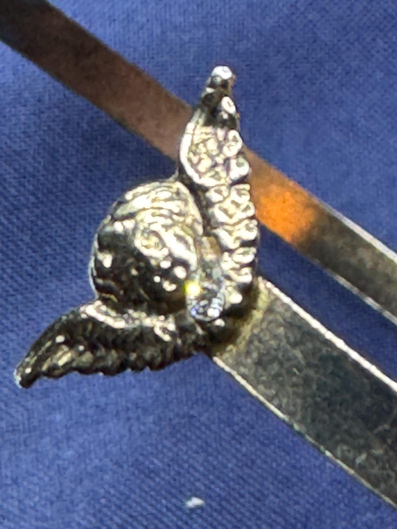 May include: Close-up of a silver-toned compass with an ornate, detailed design. The compass features a decorative element resembling a stylized bird or angel, adding a touch of elegance. The compass arms are visible, suggesting its functionality.