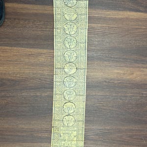 May include: A long, rectangular, cream-colored paper item with intricate, repeating circular designs and illustrations. The item is divided into sections and has a red string attached at the bottom. The item is lying on a wooden surface.