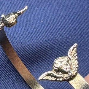 May include: A gold-toned bracelet with detailed cherub accents. The cherubs have detailed wings and are positioned on the ends of the bracelet. The bracelet is set against a blue background.