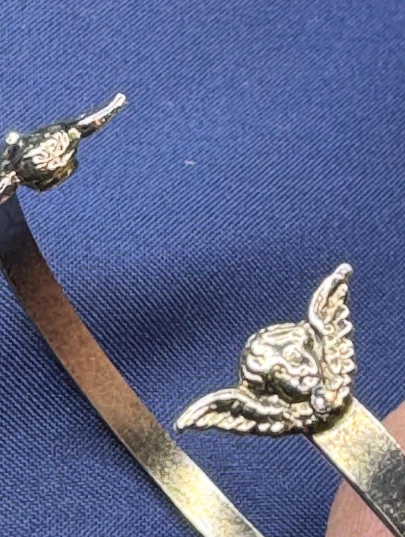May include: A gold-toned bracelet with detailed cherub accents. The cherubs have detailed wings and are positioned on the ends of the bracelet. The bracelet is set against a blue background.