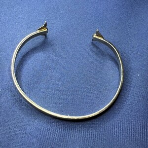 May include: A gold-colored open cuff bracelet. The bracelet has a simple, rounded band with triangular arrow-shaped ends. The bracelet is set against a dark blue background.
