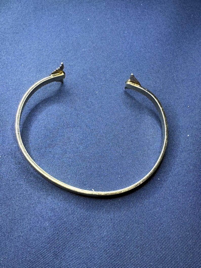 May include: A gold-colored open cuff bracelet. The bracelet has a simple, rounded band with triangular arrow-shaped ends. The bracelet is set against a dark blue background.