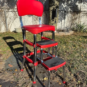 May include: A vintage red and chrome step stool chair. The chair has a rounded back, a seat, and two steps with black, textured surfaces. The legs are chrome with red accents on the feet.