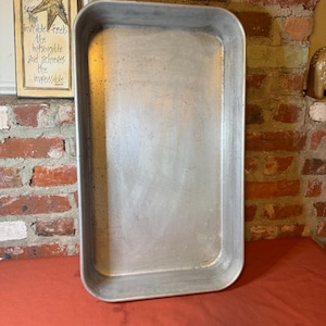 May include: A rectangular, silver-colored metal baking pan with rounded corners. The pan is empty and sits on a red surface. In the background, a brick wall and a framed sign with the word "HOPE" and a star are visible.