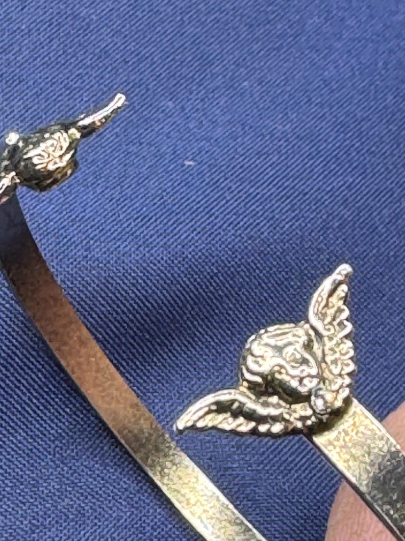 May include: A gold-colored bracelet with an angel wing design. The bracelet has a simple band with an angel figure on each end. The angel figures have detailed wings and facial features. The bracelet is shown against a blue background.