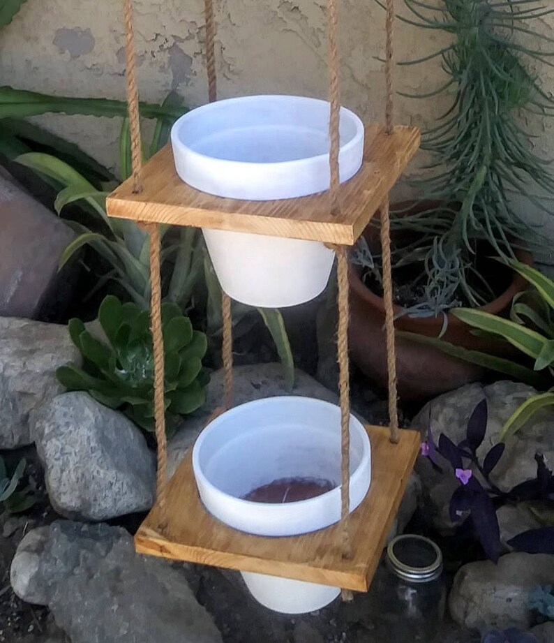 May include: Two white ceramic hanging planters with a wooden frame and rope. The planters are suspended from the ceiling.
