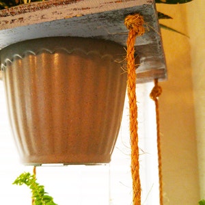 May include: A hanging planter with a light brown ceramic pot and a light brown wooden shelf. The planter is suspended by two thick, natural-colored ropes.