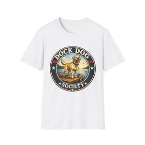 May include: A white short-sleeve t-shirt featuring a circular graphic on the chest. The design shows a yellow Labrador retriever standing on a wooden dock at sunset, surrounded by the text DOCK DOG SOCIETY in a bold, dark blue circular frame.