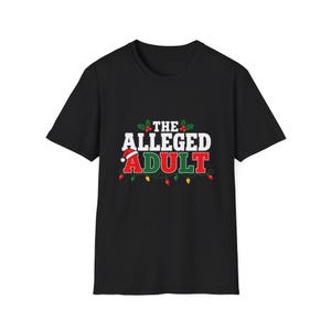 Puede incluir: Una camiseta negra de manga corta con el texto THE ALLEGED ADULT en letras blancas, rojas y verdes. El diseño incluye un pequeño gorro de Papá Noel sobre la letra A, hojas de acebo verdes con bayas rojas y una cadena de luces navideñas.